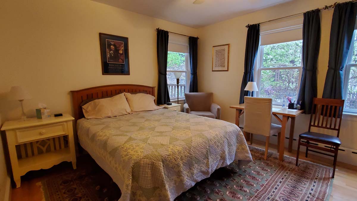 large view of room with queen bed, comfy chair, 3 windows, large nightstand/bookshelf